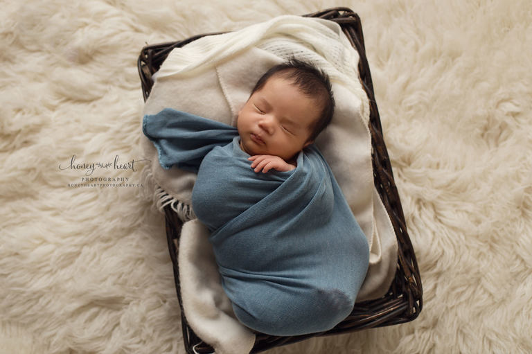 Older newborn baby pose- baby boy wrapped in blue fabric laying in a basket on cream flokati Newborn Photography Calgary