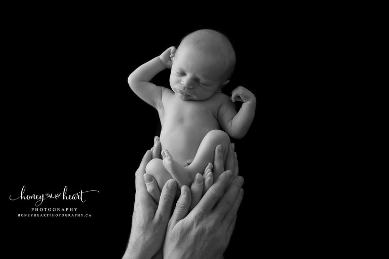 Baby boy sleeping stretched out on black backdrop with parents hands holding him Baby Photography Calgary