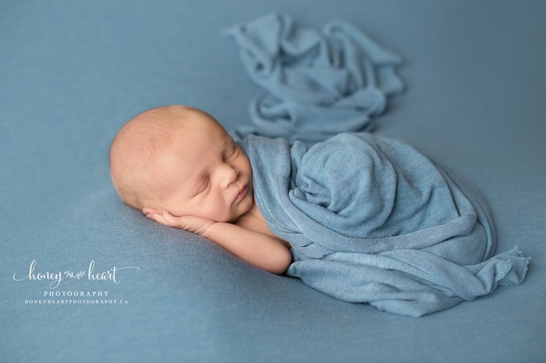 Baby boy sleeping with hand under head with blue wrap covering him Calgary Baby Photography Calgary Newborn Photography