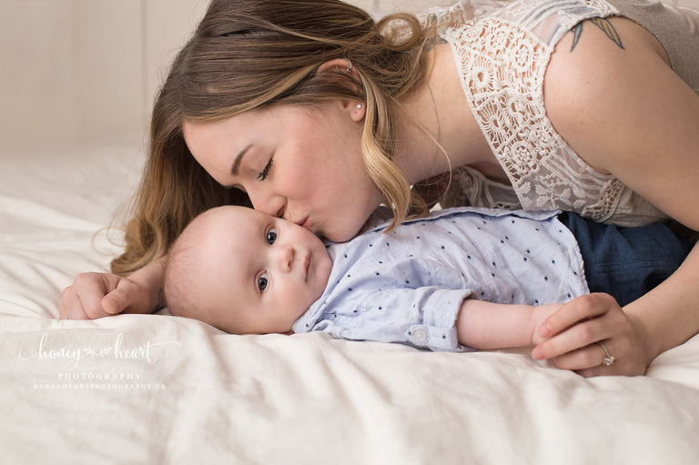 Baby boy laying on bed with mom bending down and giving baby a kiss on the cheek Baby Milestone photographers SW Calgary AB